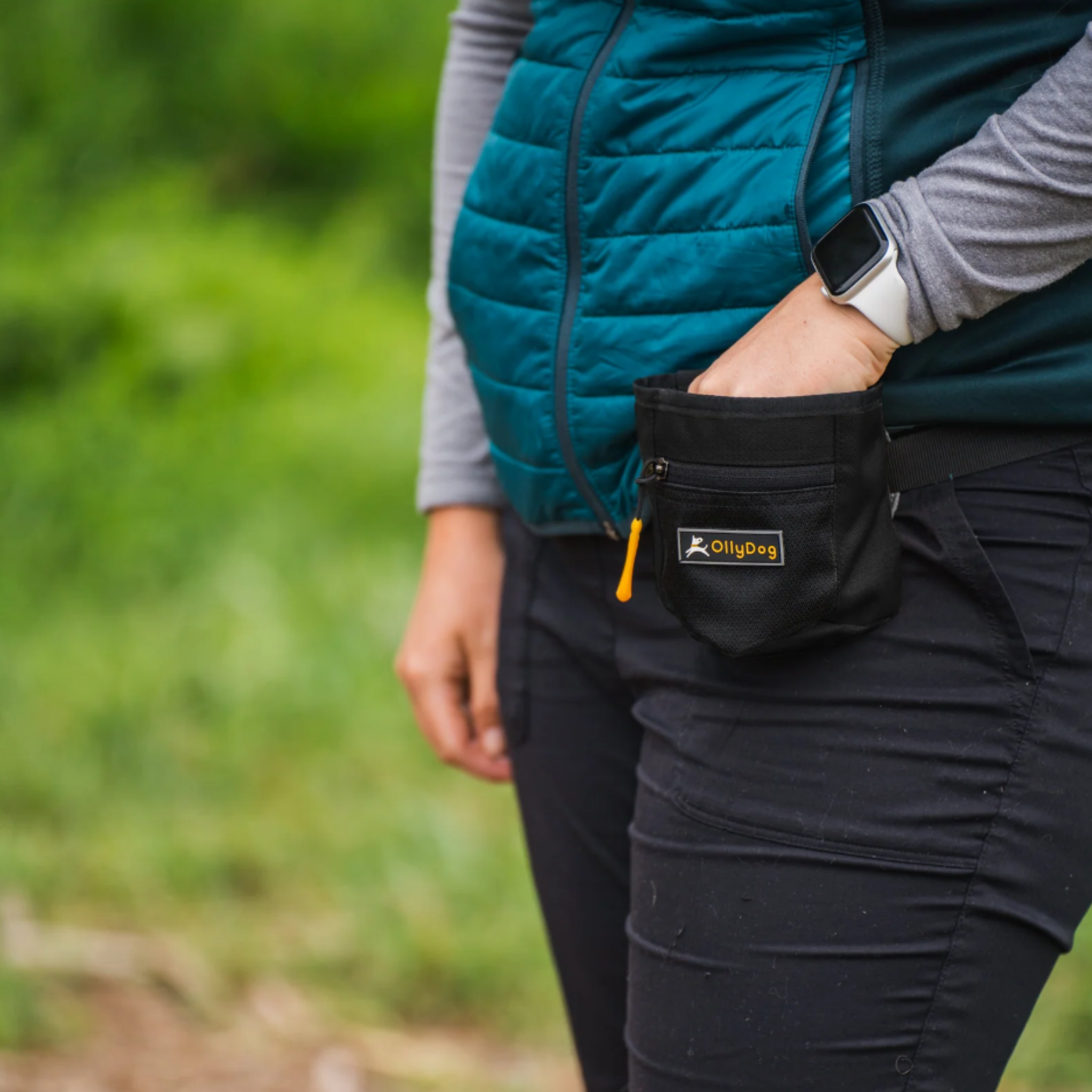 The image shows a person wearing a teal-colored puffy jacket and holding an OllyDog Goodie Treat Bag, which is a small treat pouch in a black "Raven" color, attached to their waist. The pouch is likely used to carry dog treats or other pet supplies. #color_raven