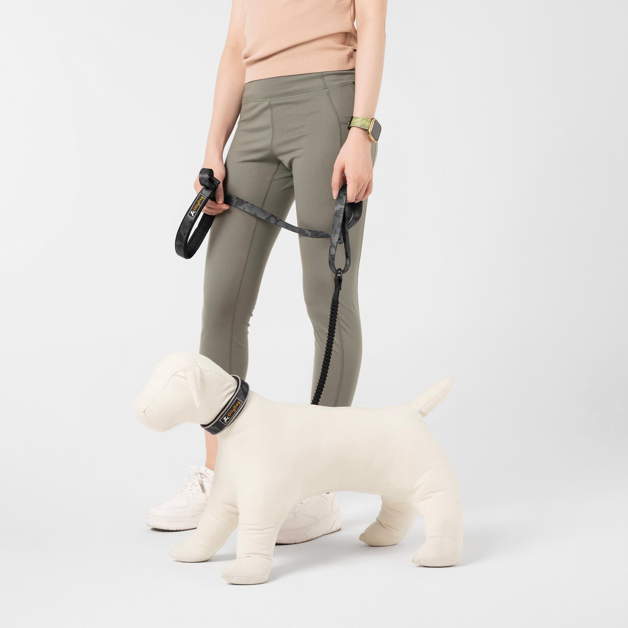 A person wearing a light-colored top and olive green leggings is holding a white mannequin dog with the OllyDog Flagstaff Adjustable Spring Leash in a dream camo (black and gray) pattern. The leash features a black padded handle for comfortable grip and a bungee section for flexible movement during dog walks #color_dream camo