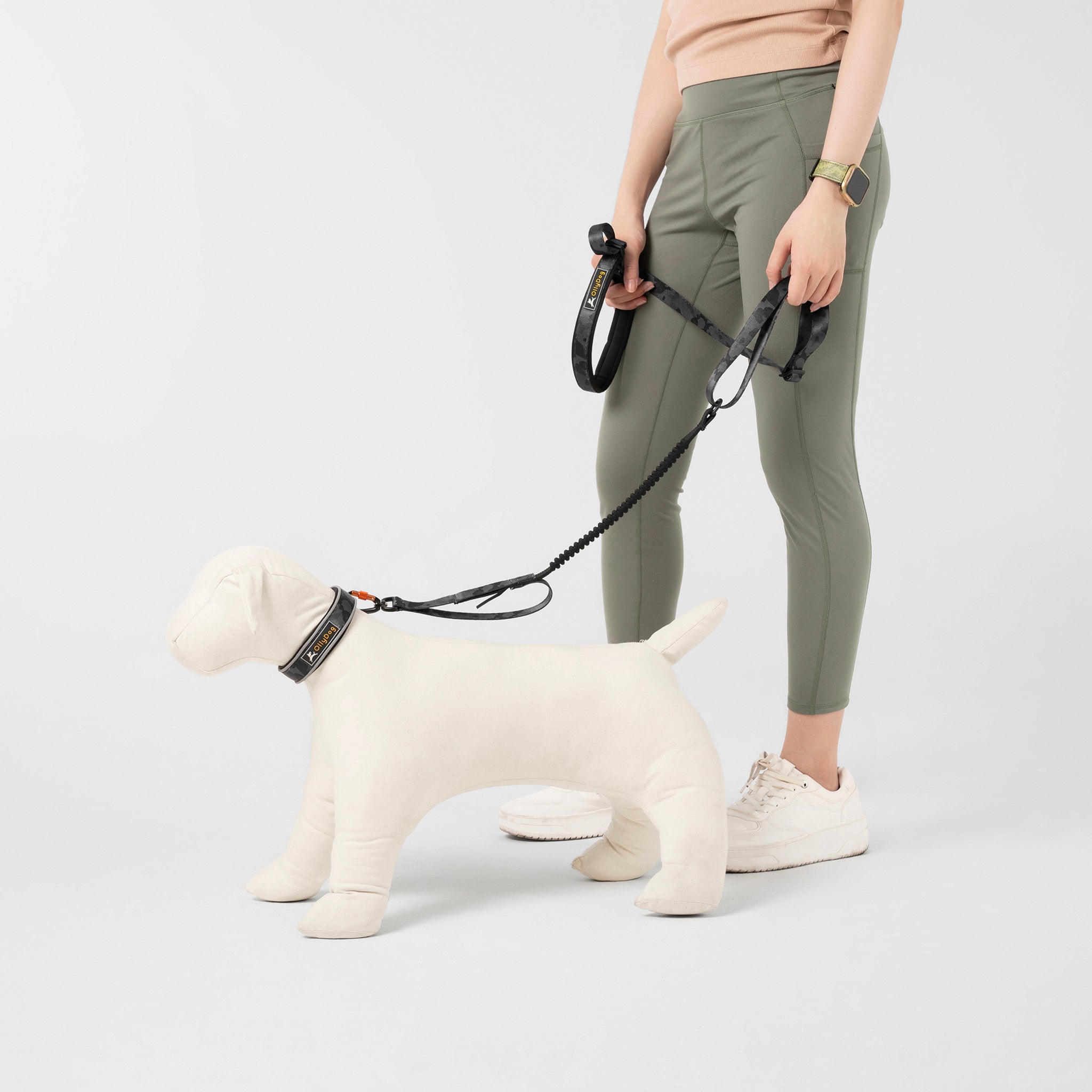 Person holding the OllyDog Flagstaff Adjustable Spring Leash in Dream Camo, attached to a dog mannequin. The leash features padded handle, waist belt, swivel carabiner, and bungee stretch for running and hands-free walking #color_dream camo