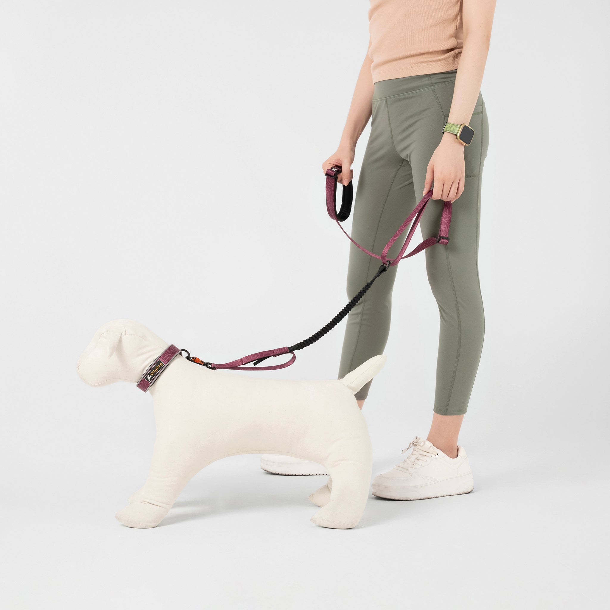 Person holding the OllyDog Flagstaff Adjustable Spring Leash in Rosewood Woodgrain, attached to a dog mannequin. The leash features a padded handle, adjustable waist belt, bungee stretch section, and swivel carabiner hook, designed for hands-free dog walking and running #color_rosewood woodgrain