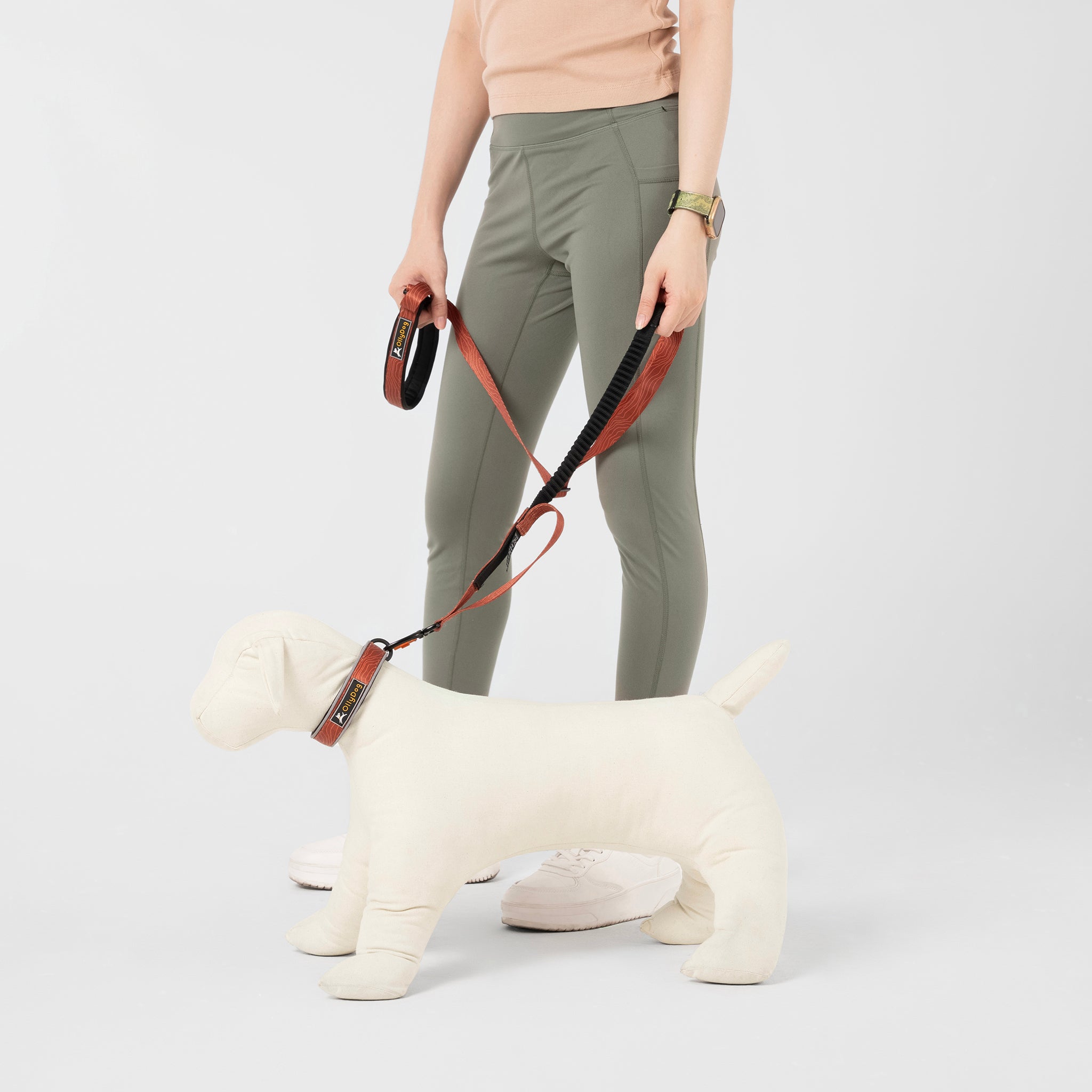 Close-up of the OllyDog Flagstaff Adjustable Spring Leash, showcasing its clay woodgrain pattern, black padded handle, bungee section for flexible walking, and a silver carabiner hook for secure attachment. This leash is designed for comfortable, hands-free dog walking and running #color_clay woodgrain