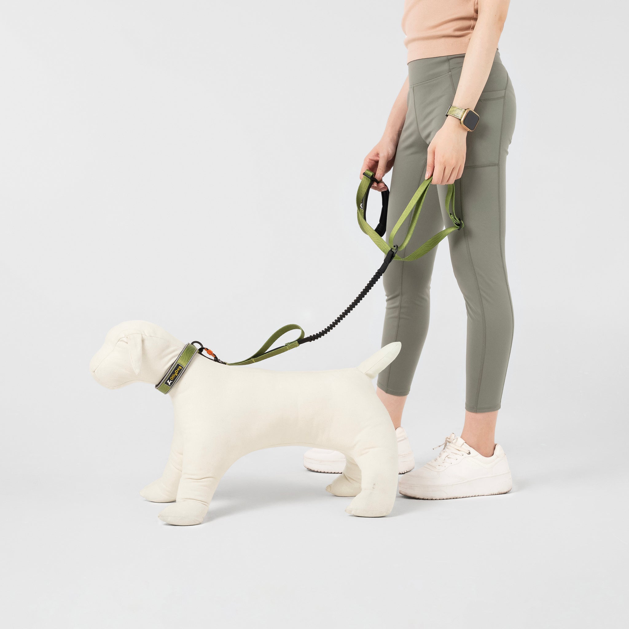 A person wearing a light-colored top and olive green leggings is holding a white mannequin dog with the OllyDog Flagstaff Adjustable Spring Leash in a cactus woodgrain (green and black) pattern. The leash features a black padded handle for comfortable grip and a bungee section for flexible movement during dog walks #color_cactus woodgrain