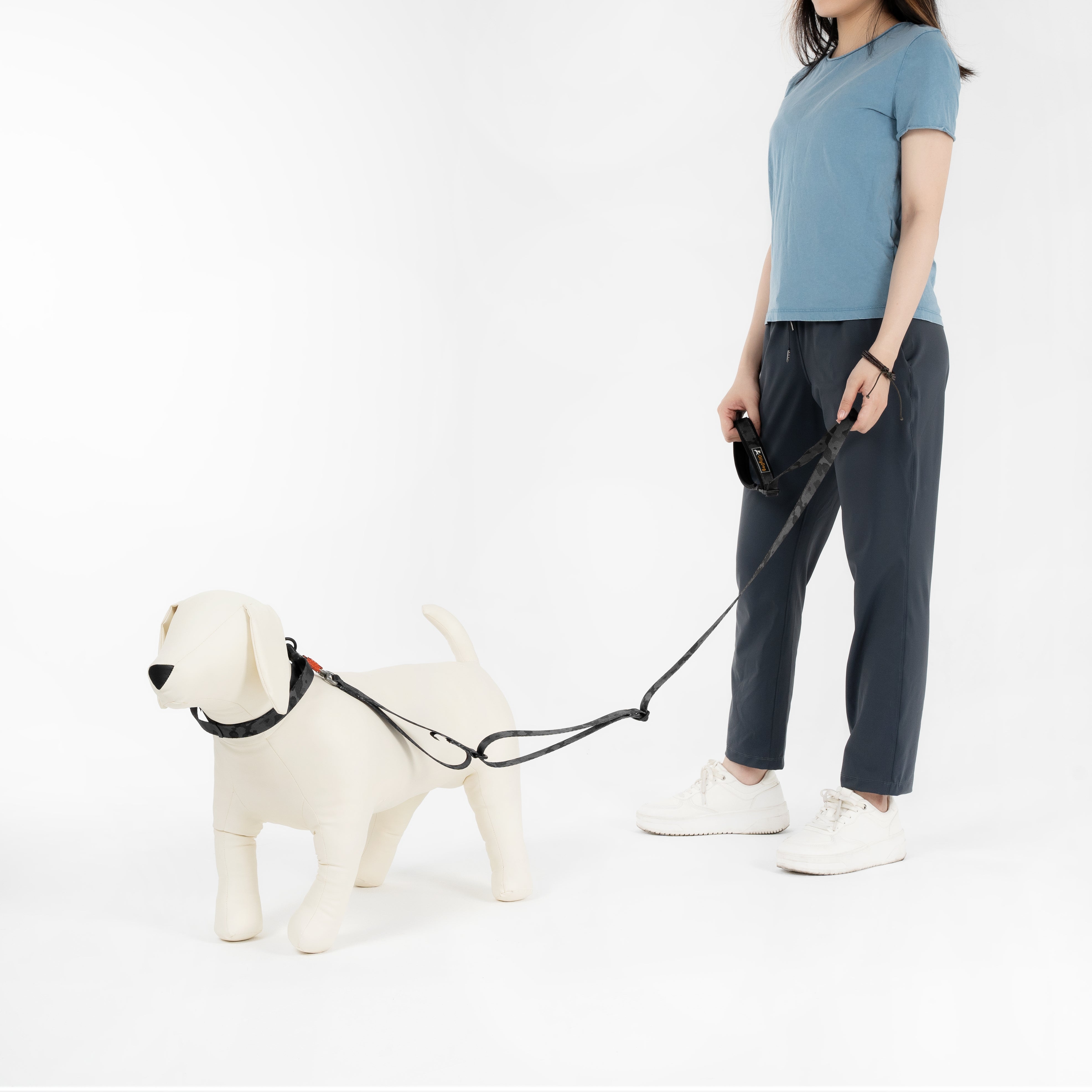 Person walking a mannequin dog wearing the OllyDog Flagstaff Reflective Dog Collar in Dream Camo, showing full-body view of leash and collar in black camouflage design, studio white background #color_dream camo