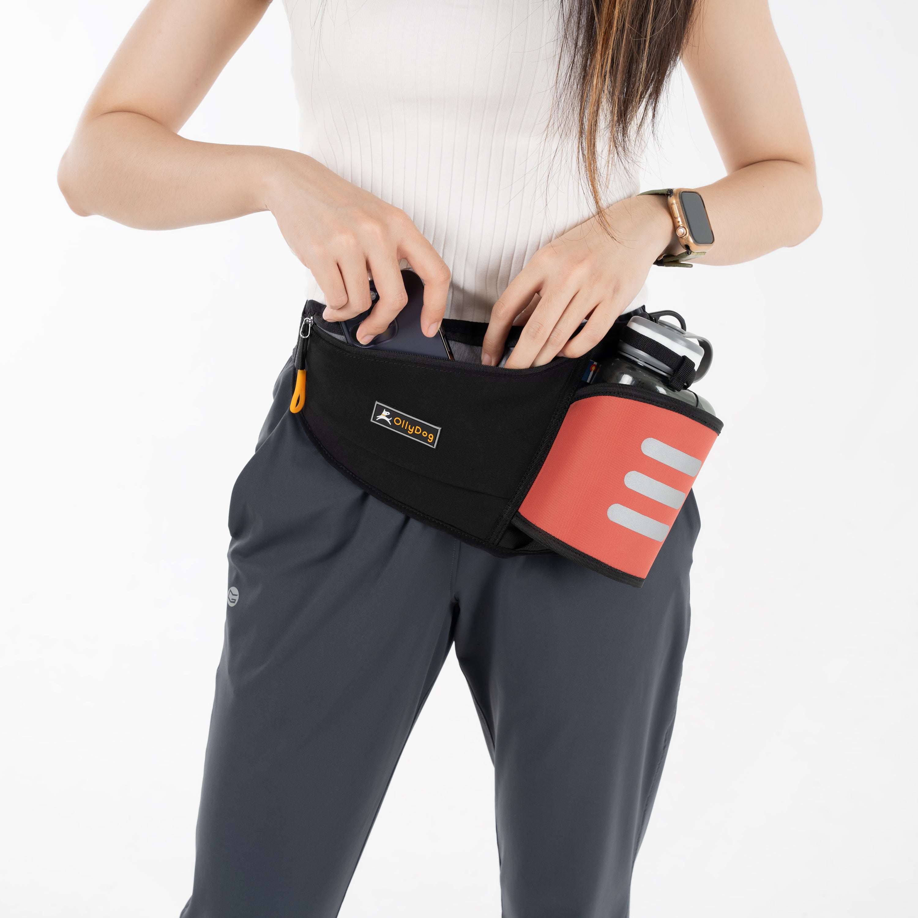 Person wearing the OllyDog Hydro-Slim Pack fanny pack in Reef color, placing items into the front zippered pocket. The pack features a reflective bottle holder with a 600mL OllyBottle, designed for hands-free convenience and outdoor activities #color_reef
