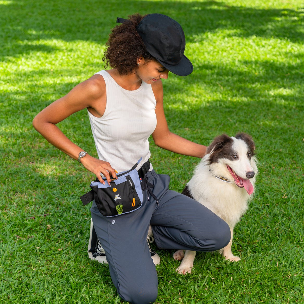 Lifestyle shot of a person wearing the OllyDog Backcountry Day Bag in blue heron color around their waist while kneeling and petting a dog on the grass outdoors. #color_blue heron