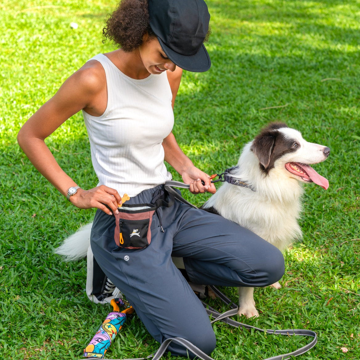 Lifestyle shot of a woman kneeling on a grassy lawn training her border collie dog while wearing the OllyDog Goodie Treat Bag in desert color around her waist and rewarding a treat. #color_desert