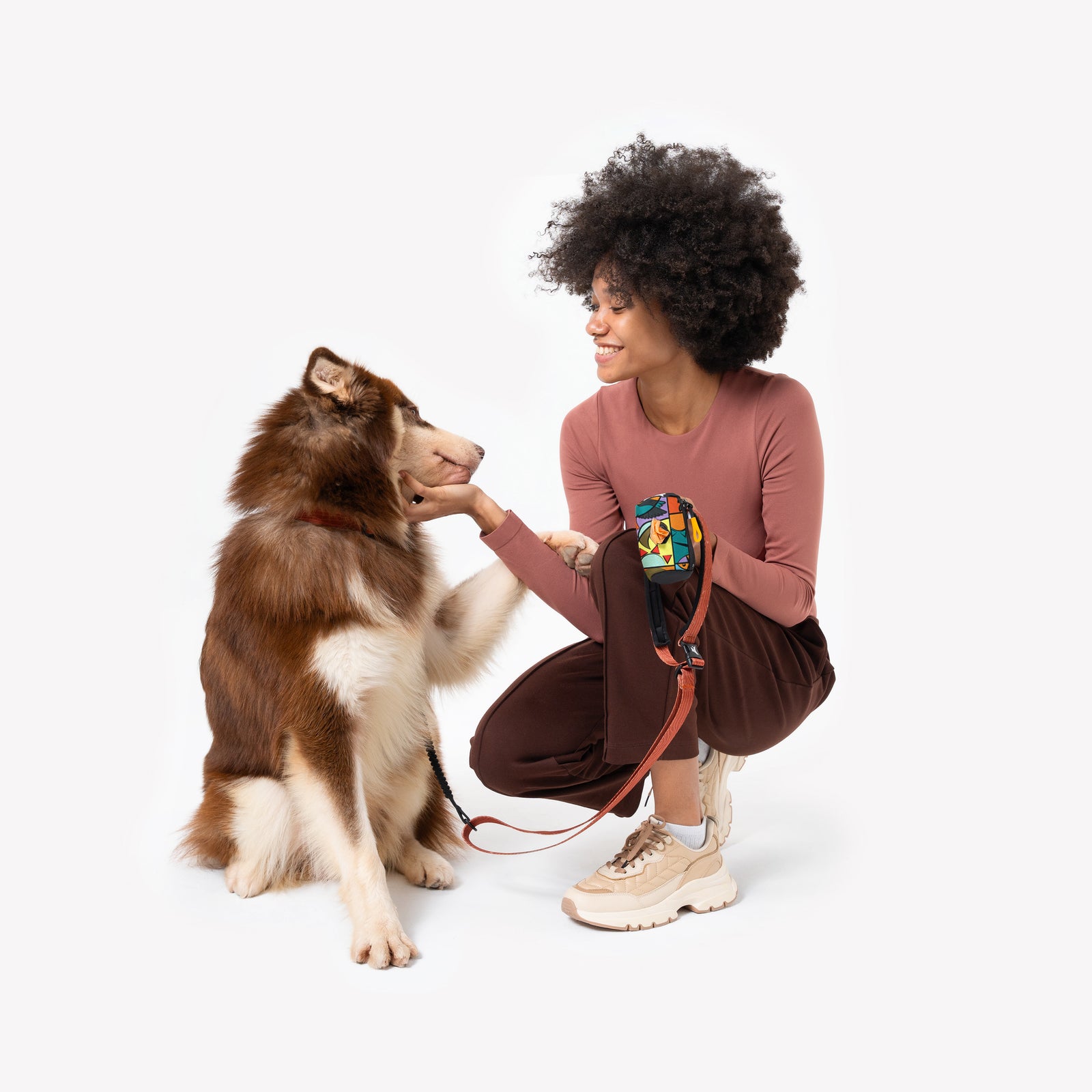 Lifestyle shot of a smiling woman crouching and petting a brown and white dog against a white background, using the OllyDog Scoop Pick Up Bag in cloud9 multi-color print attached to the leash. #color_cloud9