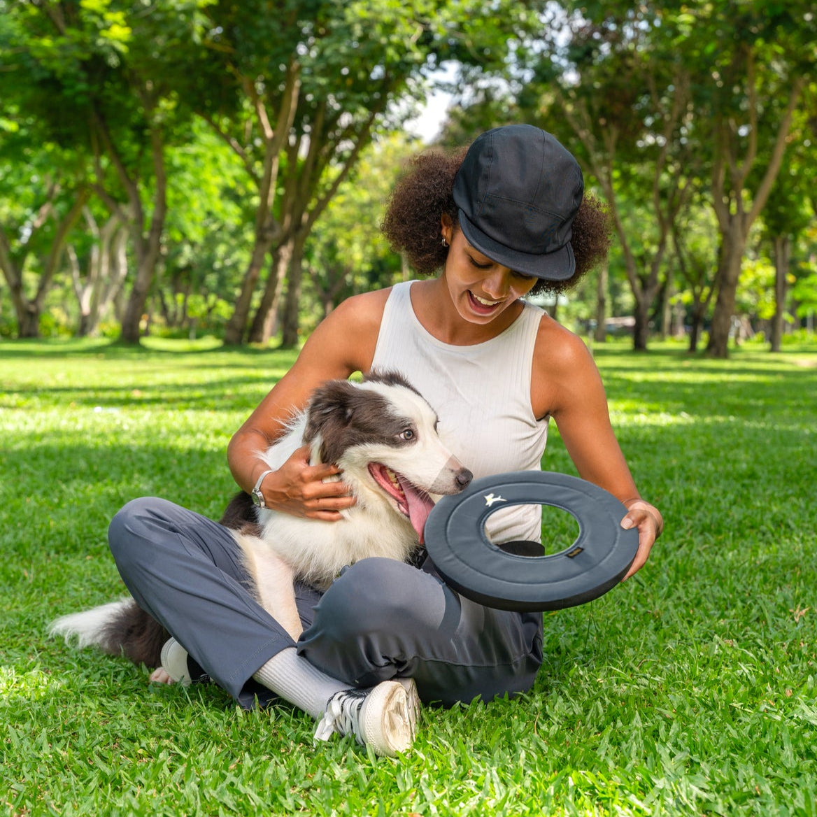 Lifestyle shot of a smiling woman sitting on a green park lawn, petting a happy border collie while holding the OllyDog Super Flyer dog flying disc in castle rock grey color.#color_castle rock
