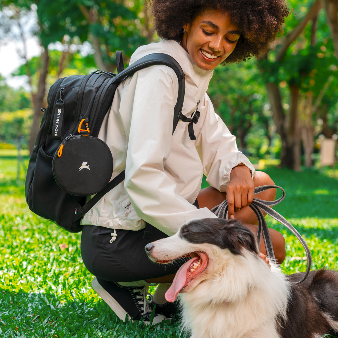Smiling woman kneeling in the park with the black backpack and attached Superbowl collapsible dog bowl, holding leash of happy black and white dog – ideal outdoor setup for pet owners and dog walkers #color_ventura '73