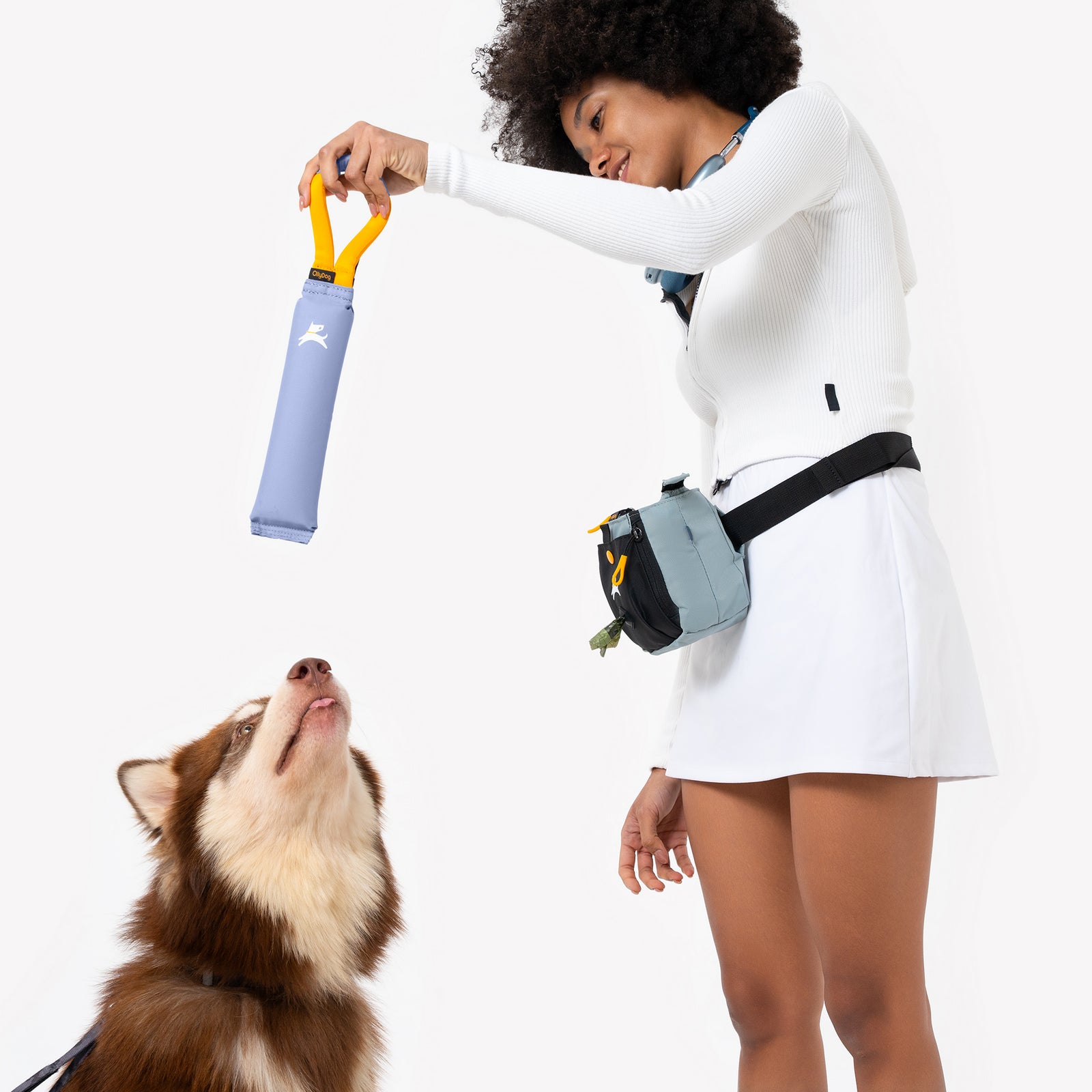 Studio lifestyle shot of a smiling woman holding the OllyDog Tugger dog toy in blue heron color above an excited dog against a white background.#color_blue heron